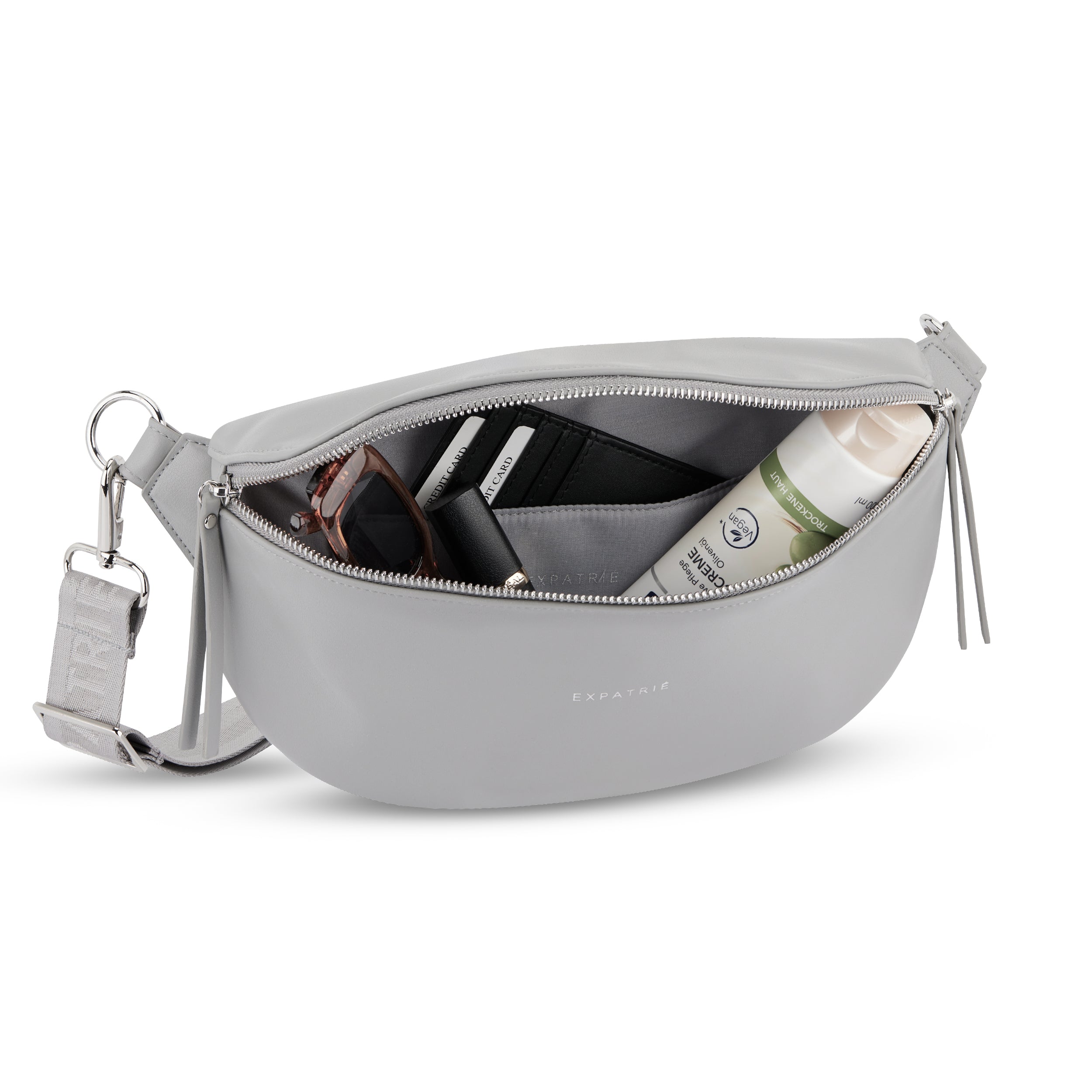 graue Bauchtasche Alice Medium von Expatrié mit geöffnetem Reißverschluss, zeigt Sonnenbrille, Lippenstift, schwarze Kartenhalter und Cremetube