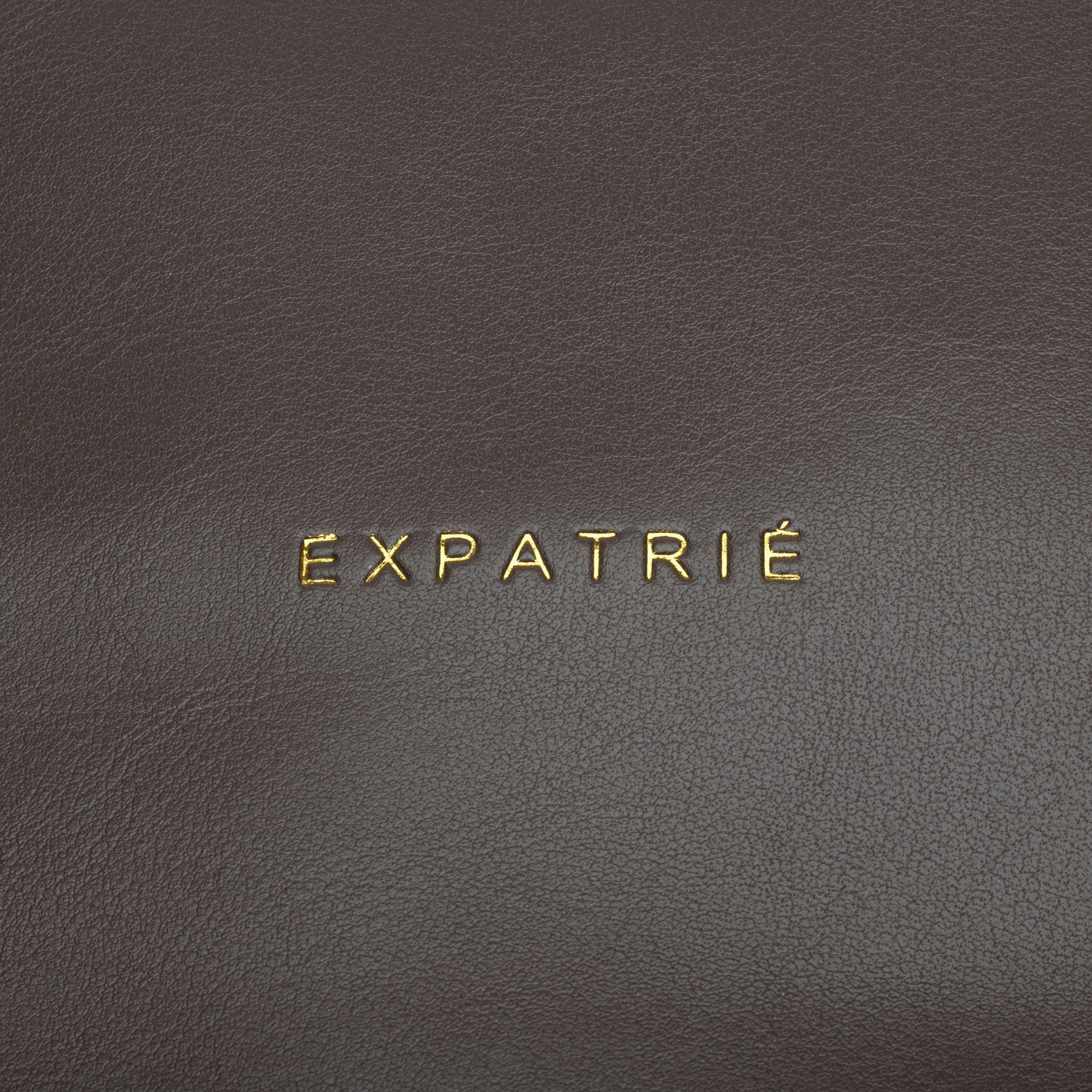 Goldene Prägung mit Schriftzug 'EXPATRIÉ' auf dunkelbraunem Kunstleder