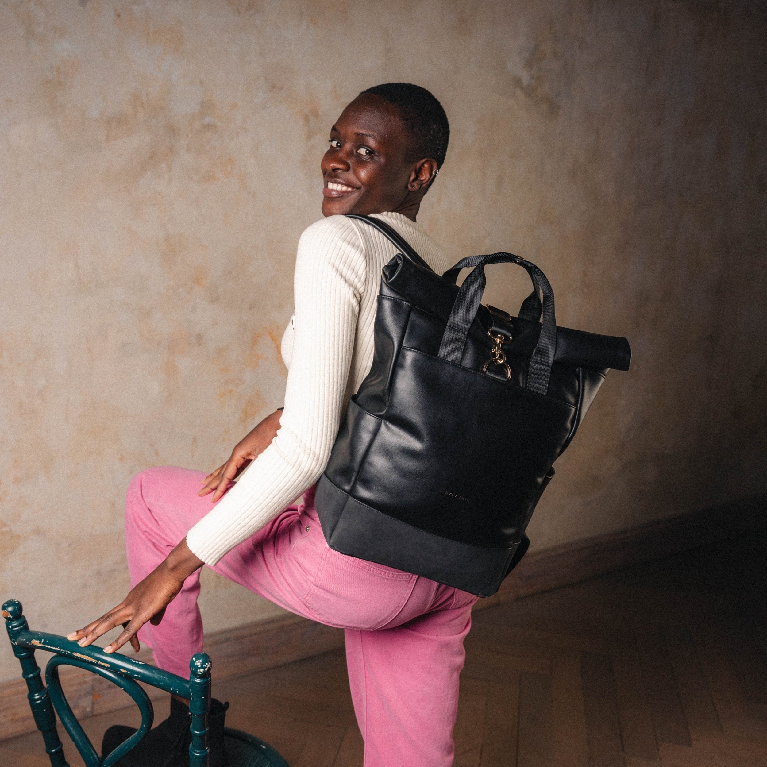 Frau mit Rolltop Rucksack Adele aus schwarzem Leder, trägt cremefarbenen Pullover und rosa Hose