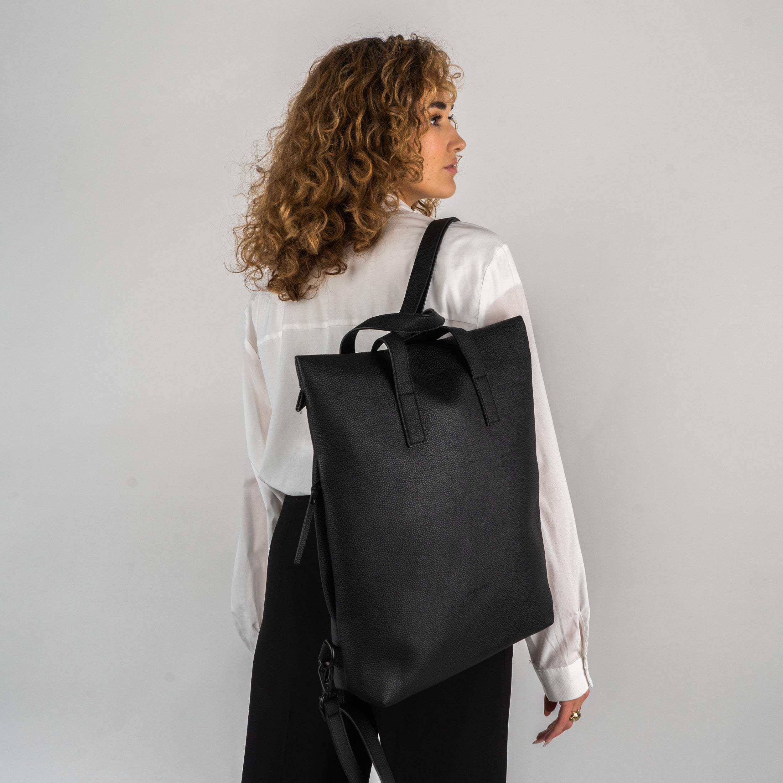 Frau mit lockigem Haar trägt schwarzen Leder-Rucksack 'Lou' über weißer Bluse und schwarzem Rock